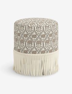 a white and brown ottoman with fringes on the bottom, in front of a gray background