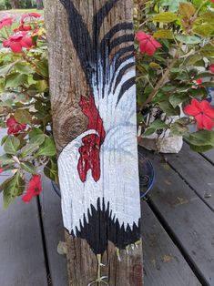 a painted rooster on a wooden post next to some red and white flowers in a pot