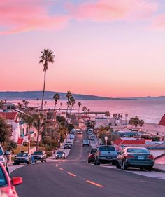 cars parked on the side of a road next to the ocean at sunset with palm trees in the foreground