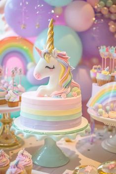 a table topped with lots of cupcakes and unicorn cake next to rainbow balloons