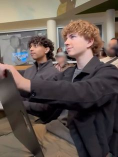 two young men standing next to each other in front of a computer monitor and another man with curly hair