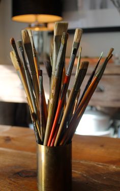 a cup filled with lots of paint brushes on top of a wooden table next to a lamp