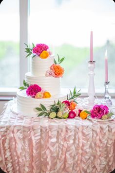 a three tiered cake with flowers and candles on the table in front of a window