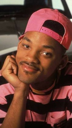 a man wearing a pink hat sitting in front of a white car with his hand on his chin