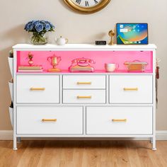 a white dresser topped with pink drawers and a clock on the wall behind it in a living room