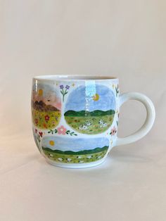 a coffee cup with flowers and mountains painted on the side, sitting on a white surface