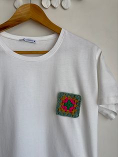 a white t - shirt with a crocheted square in the center on a wooden hanger