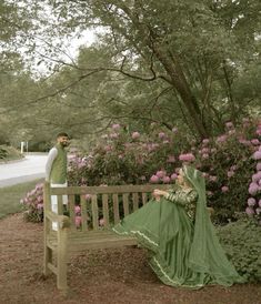 a man and woman sitting on a wooden bench in front of bushes with pink flowers