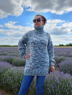 a woman standing in front of lavender flowers wearing a turtle neck sweater and blue jeans