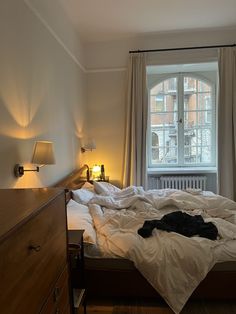 an unmade bed sitting in front of a window next to a dresser and lamp