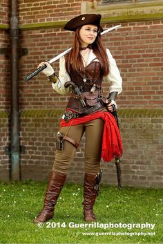 a woman dressed as a pirate holding two swords in front of a brick wall and grass