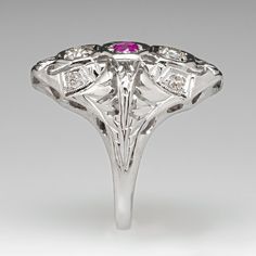 This pretty Art Deco ring features a pierced and engraved design with milgrain details and is centered with a round mixed cut ruby, weighing 0.14 carats, in a bead setting. The top of the ring is accented with two (2) bead set, round transitional brilliant cut diamonds and four (4) bead set, round single cut diamonds. The ring measures 21.0mm at the top, rises 5.4mm above the finger, tapering to 1.5mm wide and 0.6mm thick at the base of the shank. This ring is currently a size 4.5. Ruby Diamond, Art Deco Ring, Brilliant Cut Diamond, Pretty Art, Ruby, Diamond Cuts, Art Deco, White Gold, Beads