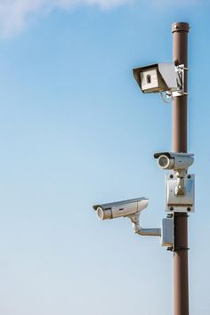 two cameras mounted to the side of a metal pole next to a street light with blue sky in the background