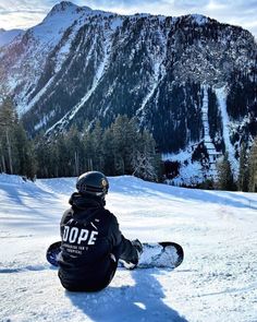 a snowboarder sitting on the ground in front of a mountain with trees and mountains behind him