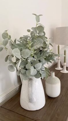 a white vase filled with greenery sitting on top of a wooden table next to a lamp