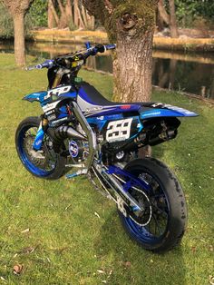 a blue and black dirt bike parked next to a tree in the grass near a body of water