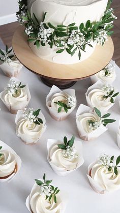 a cake with white frosting and greenery on top is surrounded by small cupcakes