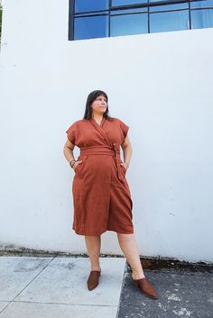 a woman standing in front of a white wall wearing a brown shirt dress and brown shoes