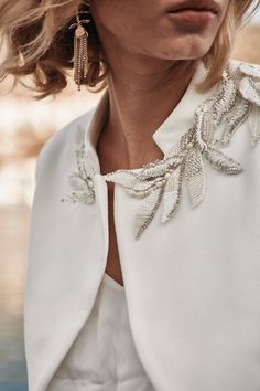 a woman wearing a white jacket and matching earrings is looking off to the side with her hand on her hip