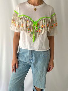 this fabulous reworked vintage festival shirt is so much fun! the white shirt is made of 100% cotton and features a crew neckline, half sleeves and a reworked raw cut cropped hem. there is fringed detailing throughout the front and the back of the shirt which features a bright neon green shoelace that ties in a bow on the centre front, real leather long fringes (around 5.5"), and colorful neon plastic beads to give it a boho festival flair. based on the tag, this shirt was presumably reworked/ha Spring Festival Tops With Beaded Fringe, Spring Music Festival Top With Fringe, Spring Fringe Tops For Music Festival, Fringe Tops For Spring Music Festival, White Cropped Festival Top, White Cropped Top For Festival, Green Spring Tops With Tassels, Spring Green Tops With Tassels, Summer Festival Green T-shirt