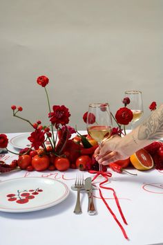 a table topped with plates and glasses filled with wine next to red flowers on top of a white table cloth