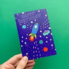 a hand holding up a birthday card with an image of a rocket ship on it