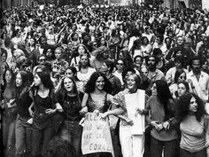 a large group of people walking down the street in black and white, with one woman holding a sign