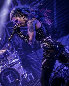 a woman with blue hair and piercings on her chest holding a microphone in front of a stage