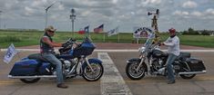 two men sitting on motorcycles in a parking lot with american flags and signs behind them