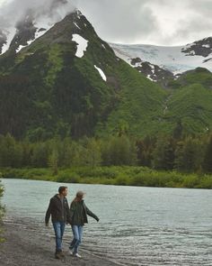 two people walking on the shore of a lake in front of snow capped mountain peaks