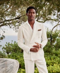 a man in a white suit standing next to a tree and bushes with his hand on his hip