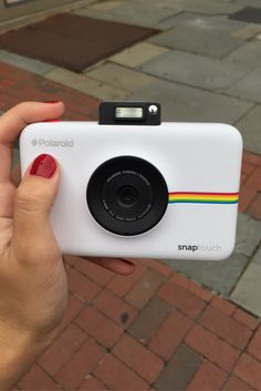 a person holding up a polaroid camera on a brick sidewalk with red nail polish