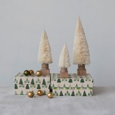 three small christmas trees sitting on top of a white box with ornaments around them and gold balls