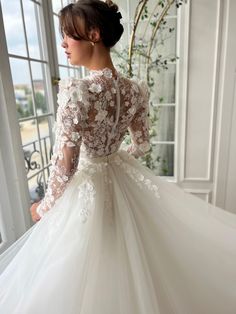 This bridal gown showcases a high neckline with a soft bow accent, paired with sheer long sleeves embellished with 3D floral appliqués. The fitted lace bodice is cinched at the waist with a delicate sash, flowing into a layered skirt featuring intricate lace beneath a tulle overlay. Ideal for weddings and formal occasions, this dress blends timeless sophistication with detailed floral artistry. Long Sleeve Dresses With Sheer Sleeves For Ceremony, Long Sleeve Tulle Ball Gown For Wedding, Long Sleeve Lace Ball Gown With Lace Bodice, Long Sleeve Gown With Lace Sleeves And Fitted Bodice, Tulle Gown With Long Lace Sleeves, Long Sleeve Lace Gown With Illusion Neckline, Wedding Night Gown With Lace Bodice And Long Sleeves, Wedding Gown With Lace Sleeves, Elegant Long Sleeve Wedding Dress With Lace Bodice