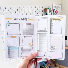 a person holding up a planner book in front of a desk with pens and pencils