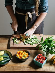 a woman cutting up vegetables on top of a wooden table next to bowls of fruit