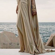 a woman in a long dress standing by the ocean with her hands on her hips