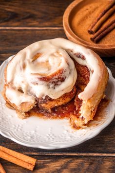 cinnamon rolls with icing on a white plate next to cinnamon sticks and a wooden bowl