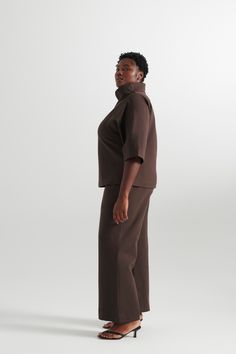 a woman standing in front of a white background wearing a brown top and wide legged pants