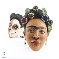 two clay masks with flowers and leaves on their heads, one wearing a skull mask