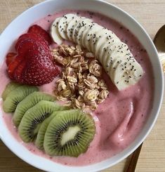 a white bowl filled with fruit and granola on top of a wooden table next to a spoon