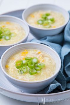 three bowls of soup with green onions and celery on a white plate next to a blue napkin