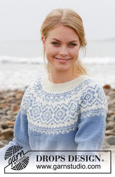 a woman standing on top of a rocky beach next to the ocean wearing a blue and white sweater