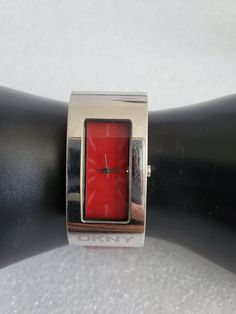 "Featuring this vintage Candy Apple Red & Silver DKNY Ladies Watch with no numbers. The bezel is silver tone color and in a rectangular shape with the letters \"DKNY\" written at the bottom. The band is wrinkle where the holes are on the shiny red part. The backsides are in a brown color with the words \"DKNY\" and \"GENUINE LEATHER\" written. A new battery was installed, and the watch runs great. Please take the time to look at the photos and video as they would describe the watch better. Watch Red Watch With Rectangular Dial For Gift, Modern Red Watch With Rectangular Dial, Modern Red Rectangular Watch, Red Watch, Red Face, Watch Gift, Vintage Candy, Candy Apple Red, Apple Red