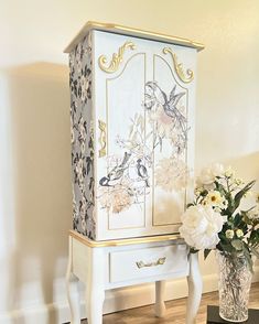 a white and gold painted armoire next to a vase with flowers in it on a wooden floor