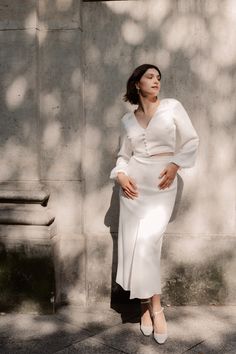 a woman is standing in front of a concrete wall wearing a white dress and heels