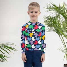 a young boy standing in front of a potted plant wearing a colorful shirt and blue pants