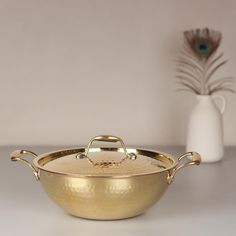 a large gold pan with handles on a white table next to a vase and peacock feather