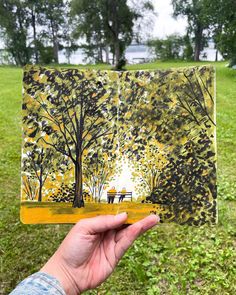 a person holding up a piece of art with trees and benches in the background on grass