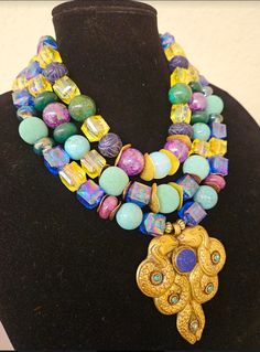 Lightweight with stunning colors, this triple strand statement necklace is destined to become one of your favorites! The featured colors are blue, yellow, purple, and green. The components include lucite cubes, magnesite, emerald, etched sapphire, mint green designer beads, white moonstone and copper and gold tone wavy metal spacers. The pendant is a hand crafted Tibetan brass repousse double dragon with inlaid lapis lazuli and turquoise. Adjusts from 17-20" and I can add additional chain links if requested. Closes with a gold tone lobster claw clasp and a 3" extender chain. From the Atelier of Kat Kouture! A perfect size so as not to overwhelm a petite woman. One Of A Kind Multicolor Fusion Jewelry, Multi-strand Czech Glass Jewelry With Large Beads, Unique Multi-strand Jewelry With Colorful Beads, Czech Glass Large Beads Multi-strand Jewelry, Unique Multi-strand Gemstone Beads, Spiritual Multi-strand Necklace With Unique Variations, Multicolor Multi-strand Spiritual Beaded Necklaces, Unique Multi-strand Large Beads Jewelry, Unique Multi-strand Jewelry With Large Beads